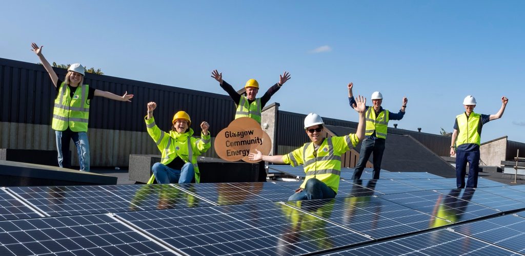 Volunteers from Glasgow Community Energy celebrate with arms raised at the site of their new solar array on the roof of a secondary school