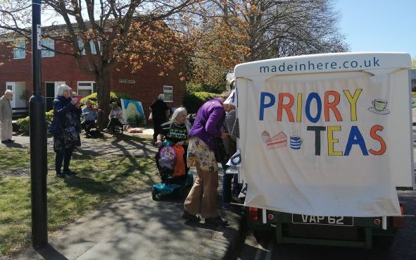 Image shows local neighbours visiting a milk cart with a homemade banner than reads 'Priory teas'
