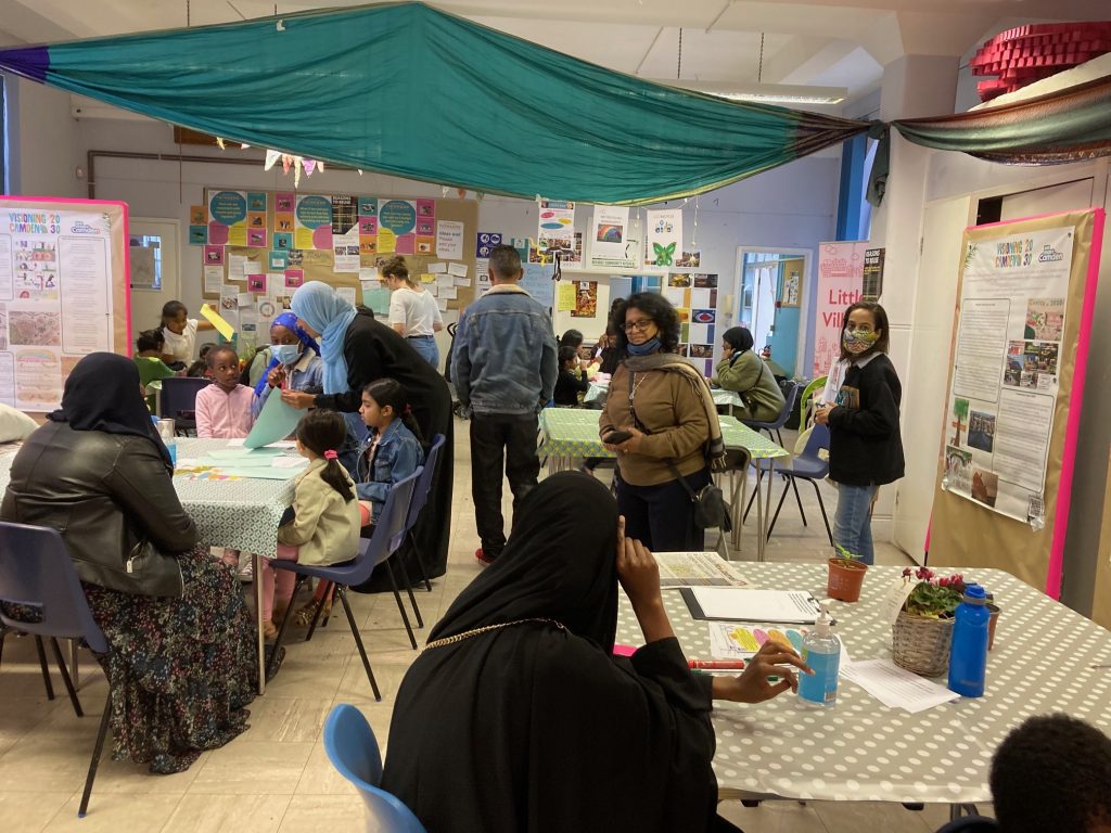 Interior view of Think & Do space, where people are gathered around tables to talk, enjoy tea and cake and take part in the many activities and discussions. In the background are 'ideas walls', as shown in the next picture. 