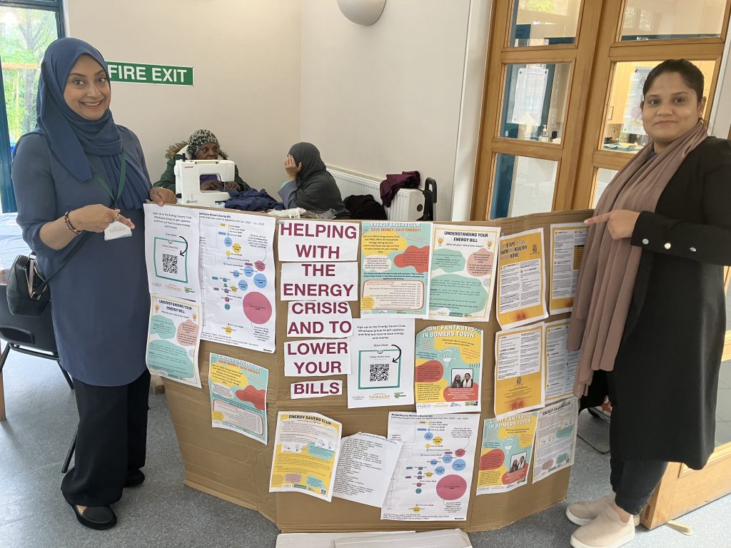 Two women, one wearing a head scarf, stand on either side of an information board entitled 'Helping with the energy crisis to lower your bills'. 