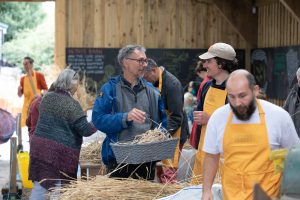 A large barn is the setting for Sheffield's threshing day. A grower holds a basket of wheat, he's in discussion with a volunteer. Various other people mill around. 