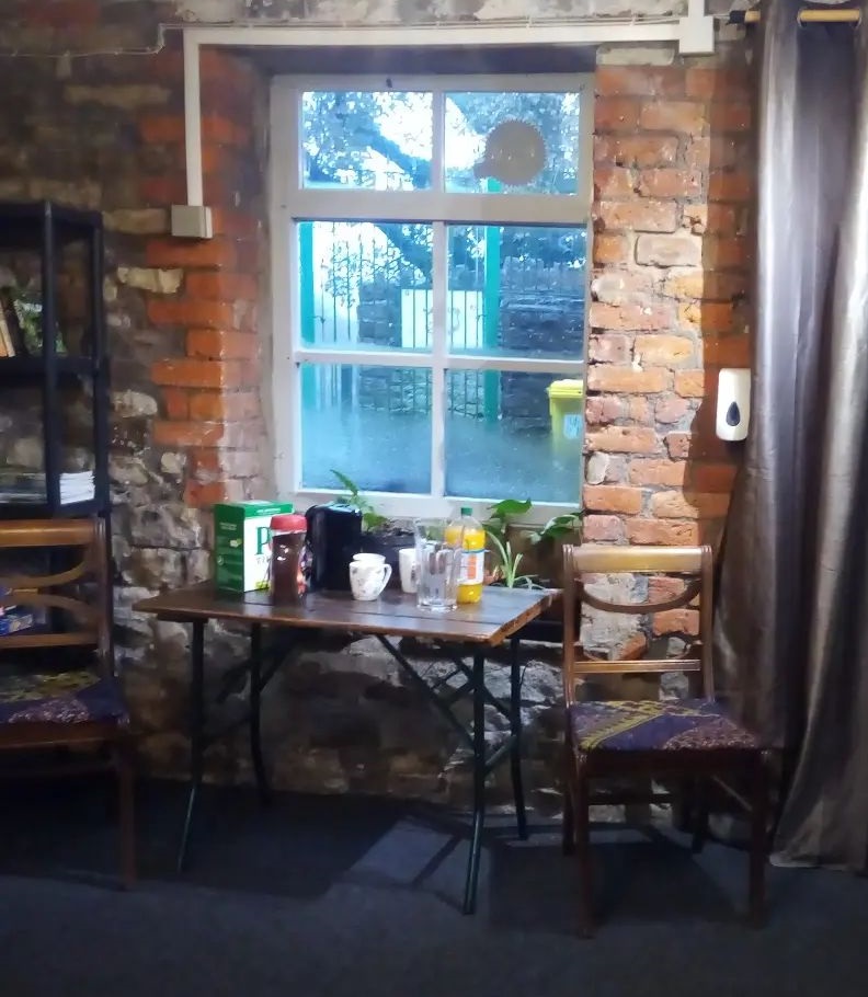 Chairs and a small table stocked with drinks and snacks make up Transition New Mills welcoming space, part of a network across the town. A bright window with cosy curtains make up the backdrop, nearby there is a bookcase with books and newspapers to browse and board games to play. 