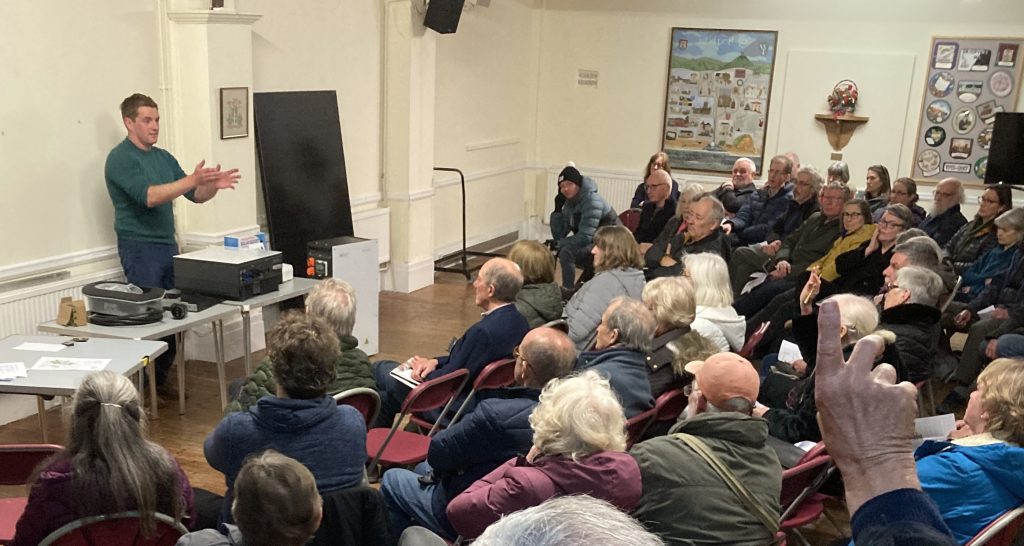Residents of Bridport gather in a community hall to hear about solar power. A speaker stands at the front of a packed semicircle of chairs to address the meeting. 