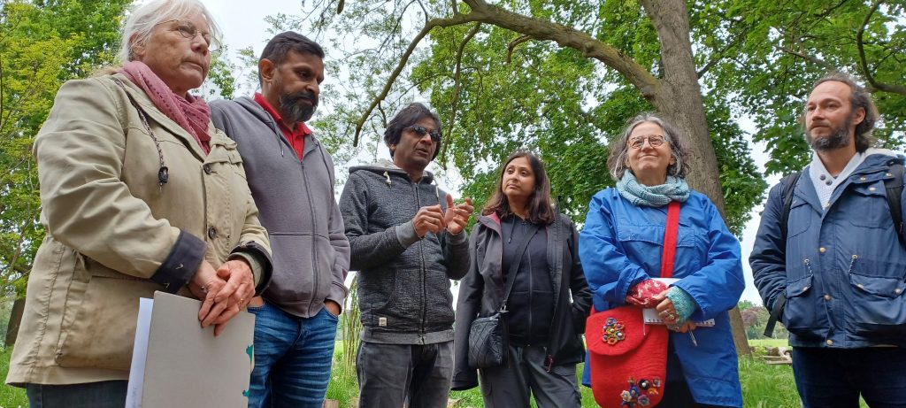 Six people stand in a row, telling the visiting group about Transition Town Ilford's many projects. Behind them are mature trees and lush, long grasses of their Forest Garden on the edge of a public park. 