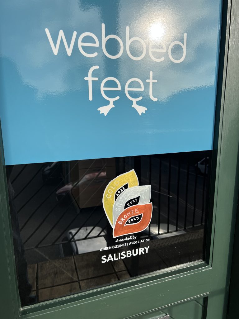 A shop window showing the business award logo, with three leaves in gold, silver and bronze. The upper part of the window has a blue sign with the name of the business Webbed Feet (each 'e' has a little foot sticking out underneath).  