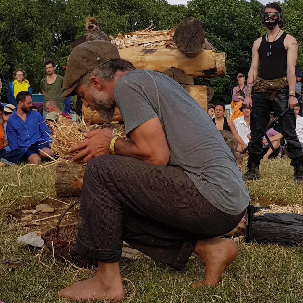 A black man in bare feet, woodworker Richard Anderson kneels to light the closing campfire as people sit around it in the background. 