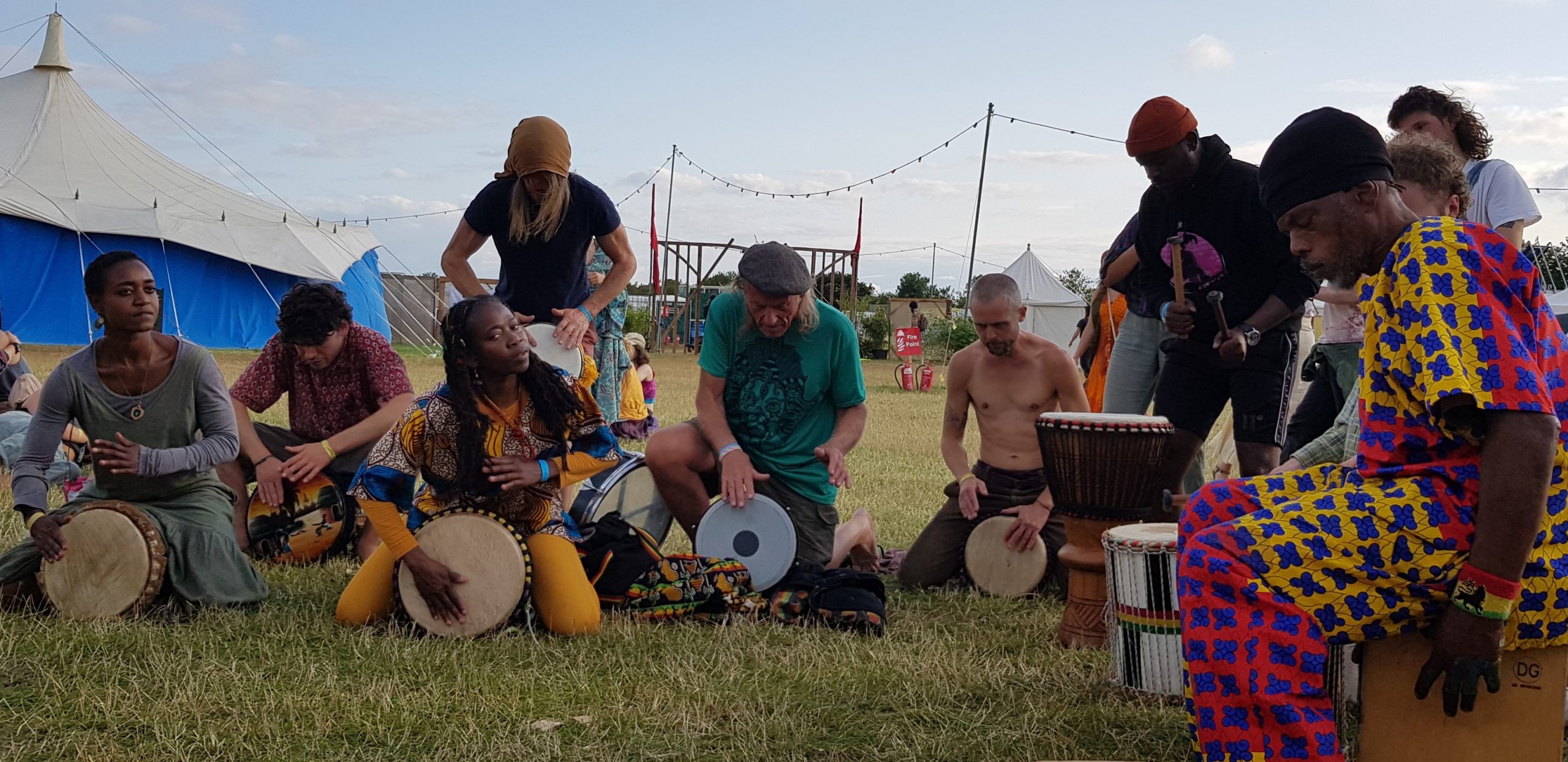 A group of pepole sitting in the grass in the evening light, playing a variety of drums. Behind them are the tents and bunting of the fair's main field. 