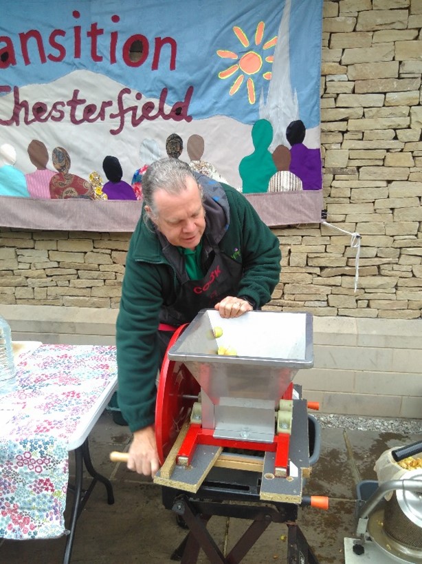 A person in a green fleece turns the handle on the apple press to turn the fruit to juice. A colourful handmade Transition Chesterfield banner hangs on the wall behind. 