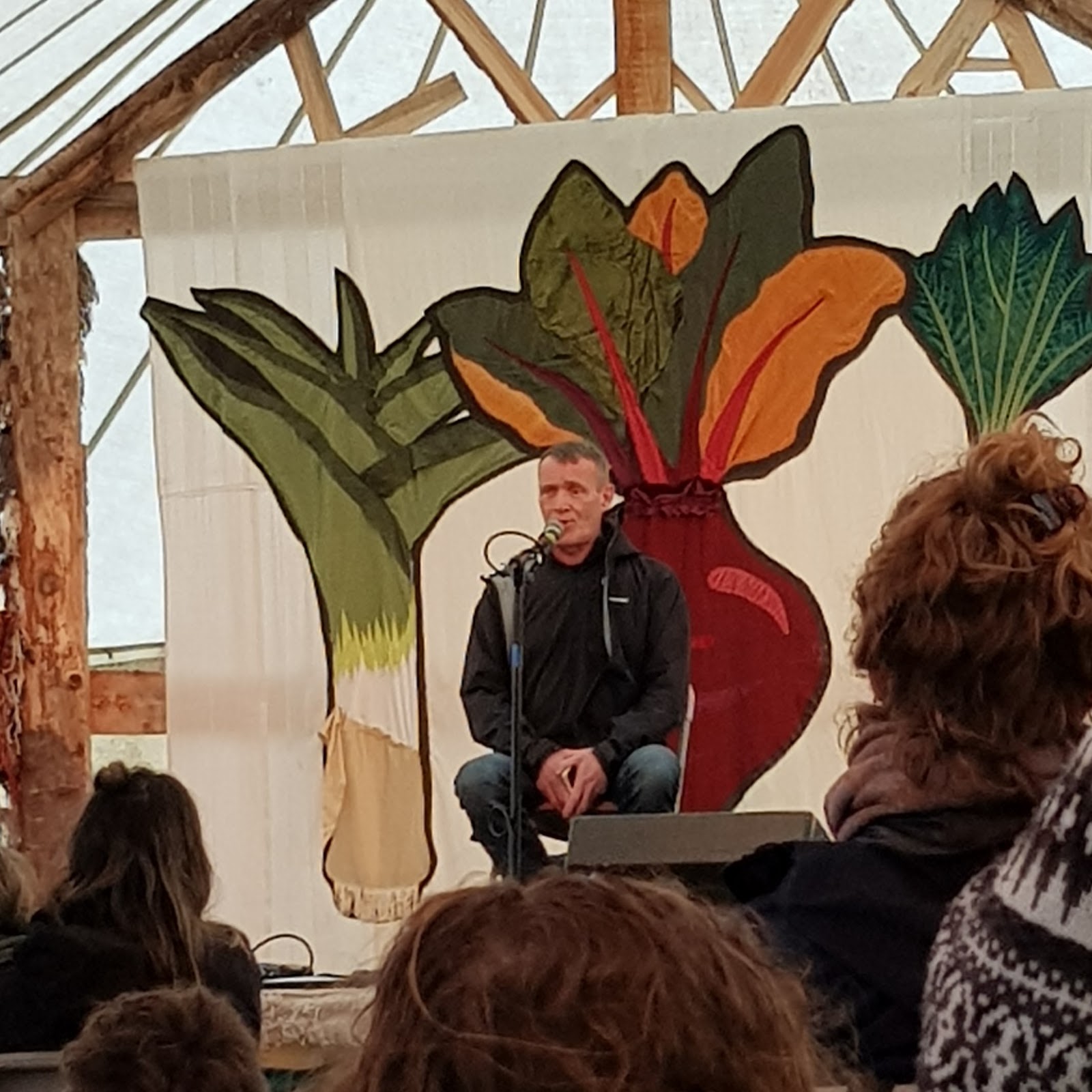 A man, Thomas McCarthy, sits on a stage speaking into a microphone. Behind him is a mural of a leek, a beetroot and a lefty veg. 