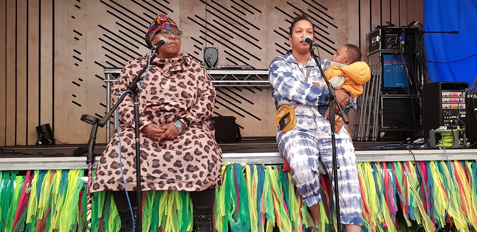 Two black women sit on the edge of a stage decorated with colourful streamers, one cradles a baby as she speaks to the workshop - and he is captivated! 