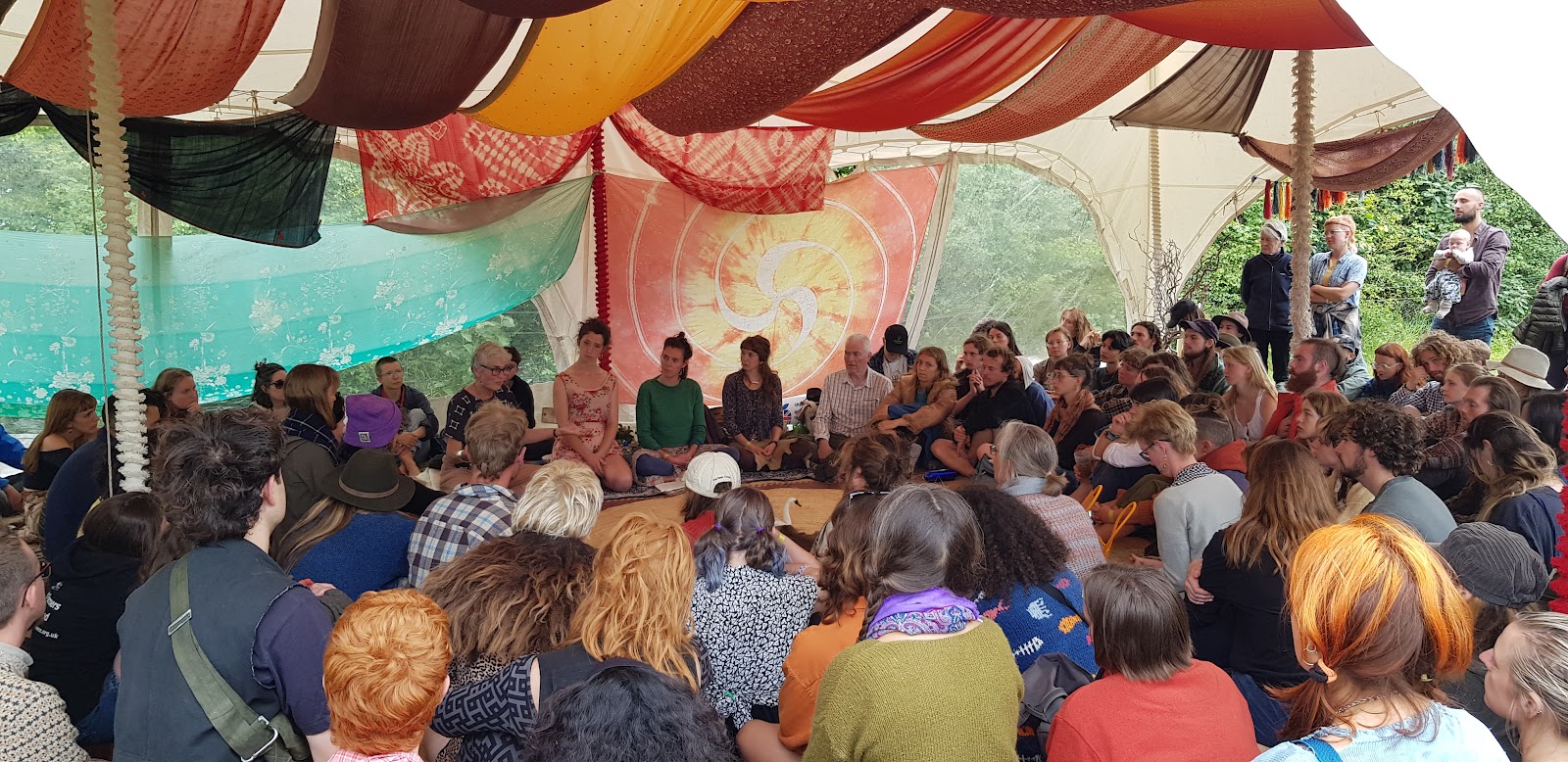 A colourful tent decorated with tie dye scarfs is packed out for the workshop, with some participants sitting on the ground and others standing around the edge. 