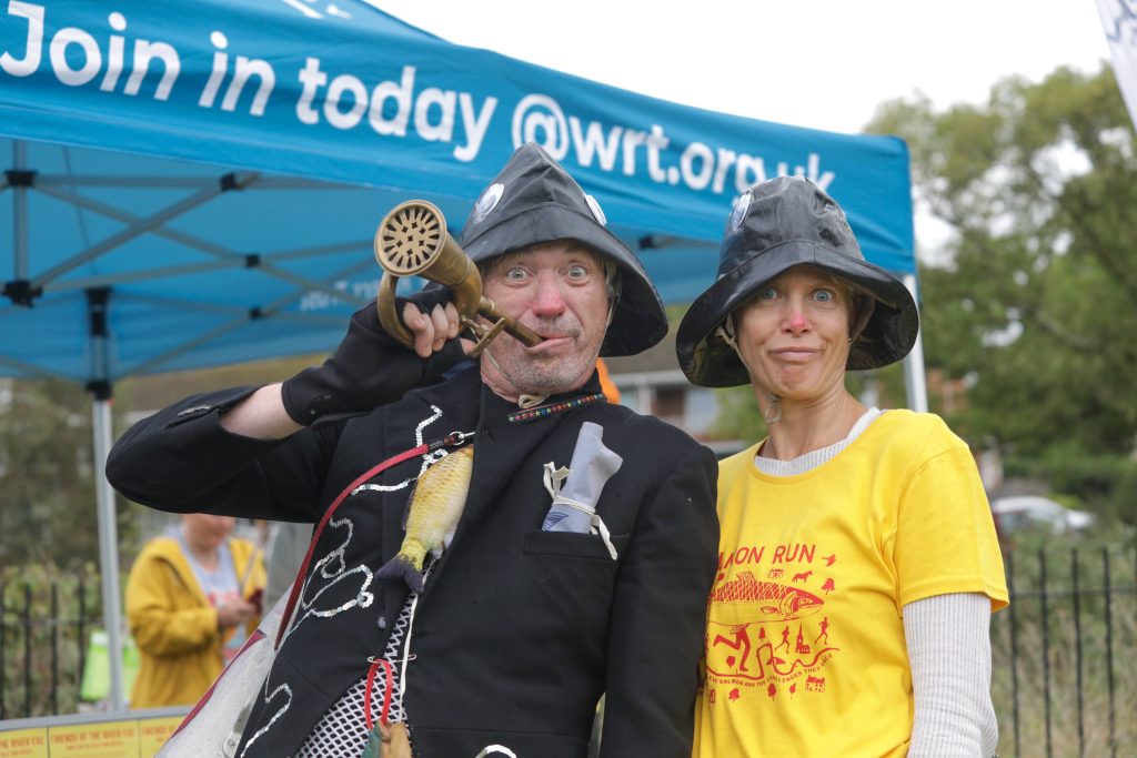 Two people stand in front of a blue gazebo,  a man in a black coat with the route of the salmon migration embroidered on it, a souwester hat and a strange brass trumpet. A woman in a yellow salmon Run stands beside him, also wearing a souwester, with a quirky look on her face... 