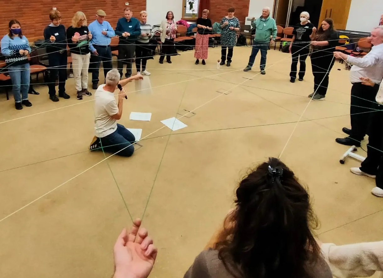 A circle of people stand in a large room, one person in the centre is holding a piece of string which is connecting members of the circle.