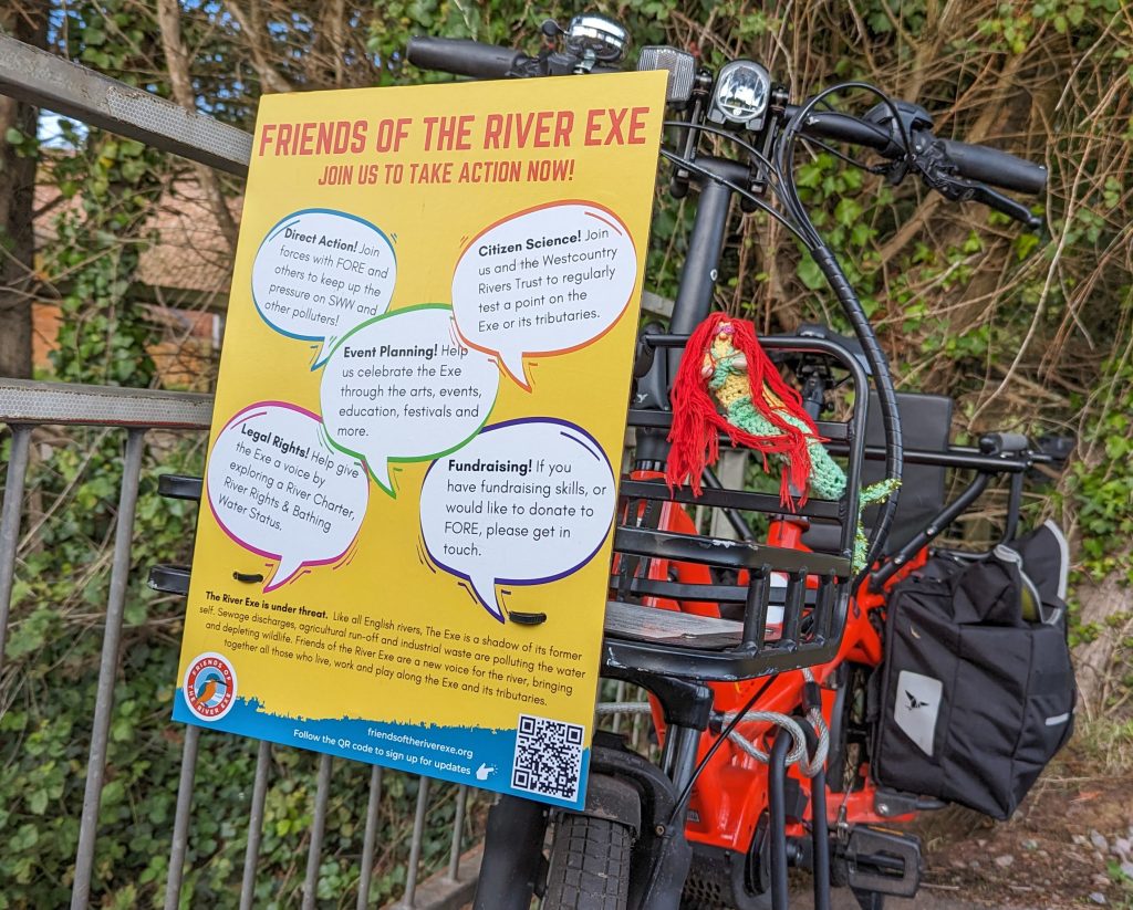 A red Tern cargo bike has a poster for 'Friends of the River Exe' attached to its front basket, inviting people to join in with Direct Action, Citizen Science, Event planning, fighting for Legal Rights and fundraising. A crochet mermaid with red hair sits next to it. 