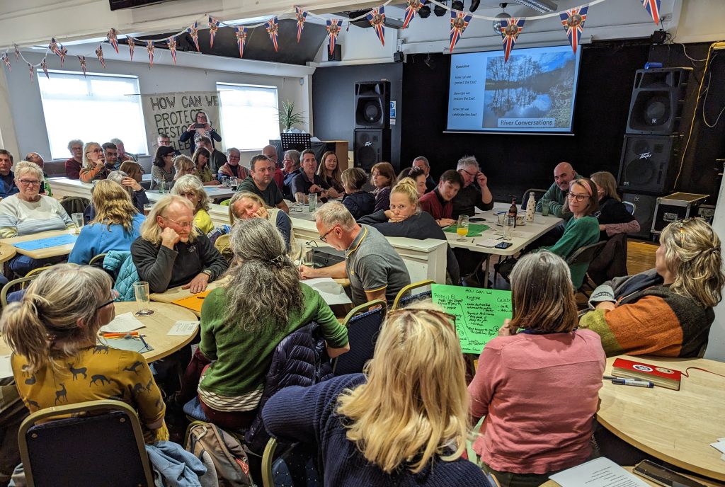 A packed room with people at tables watch a presentation about the Friends of hte River Exe project, and discuss and write down ideas that the community could take forward toenhance the river. 