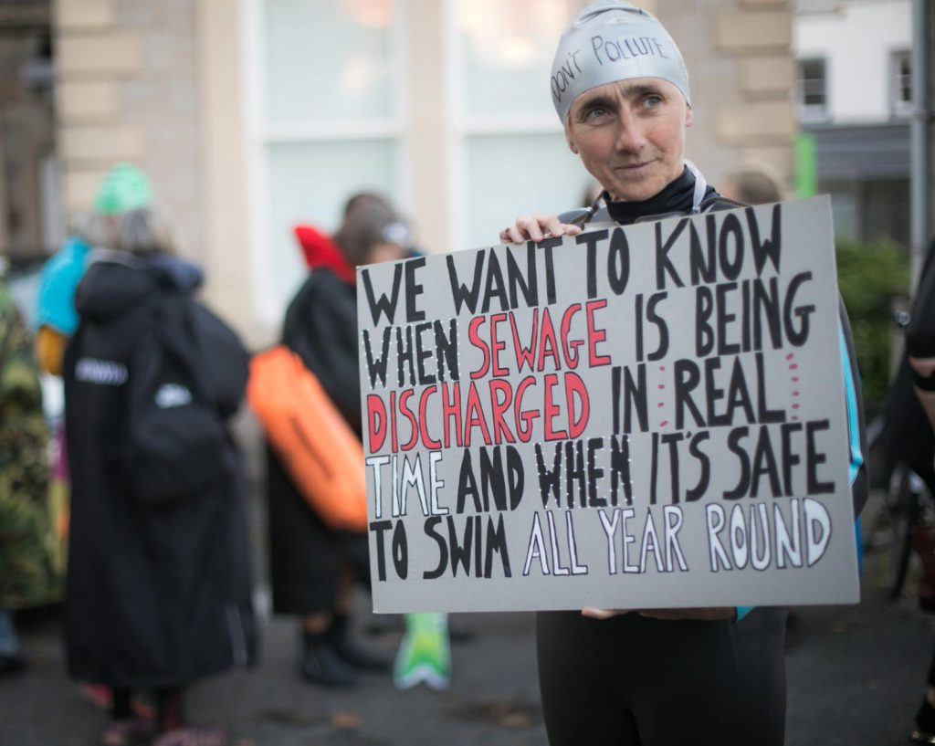 A protester in a wetsuit holds a cardboard sign which reads "We want to know when sewage is being discharged in real time and when its safe to swim all year round." In the background, other protesters are out of focus, but swim robes and bright orange tow floats can be seen. 