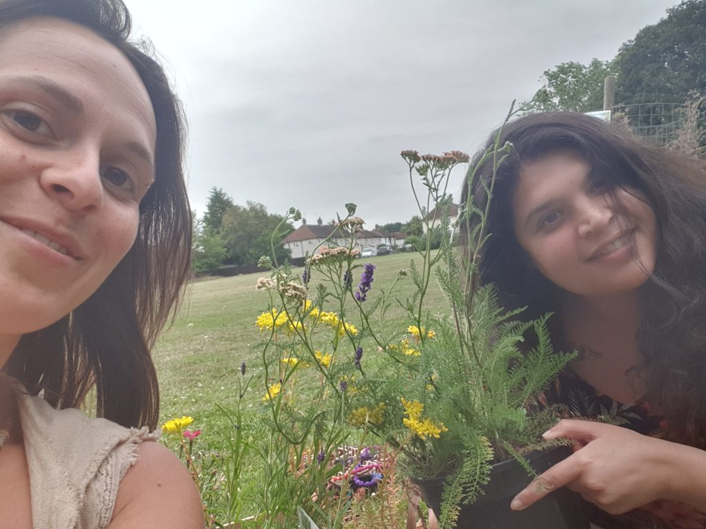 Two women with wildflowers and other plants in front of a green space in the village. 