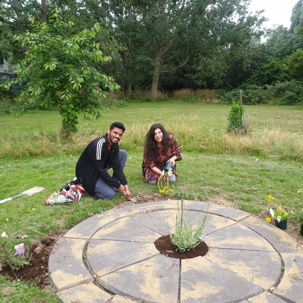 A man kneels and uses a trowel to dig a hole for a new plant, which a woman next to him is tipping out of its pot. In front of them is a circle of slabs which will soon be surrounded by flowers and plants. Behind them, young trees can be seen as well as areas of long and short grasses. 