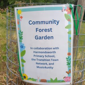 A laminated sign attached to a tree guard reads "Community Forest Garden in collaboration with Harmondsworth Primary School, the Transition Town Network and Music4Unity". 