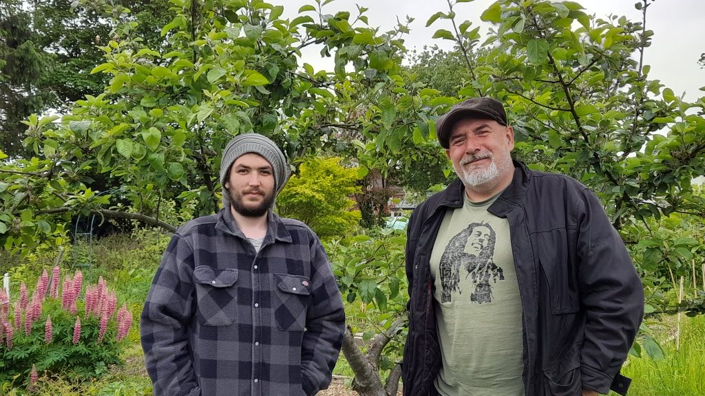 Two men stand, Ben in a woolly hat and checked shirt and Steve in a flat cap and Bob Marley t-shirt, stand in front of a community garden with apple tree, shrubs and flowers. 