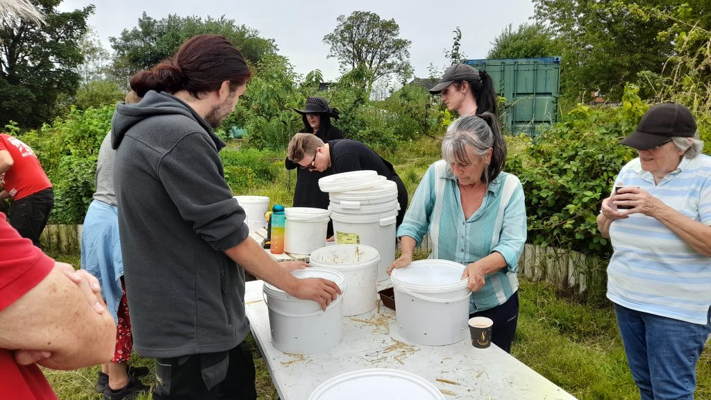 Participants of all ages gather around a table out of doors with large white catering tubs. In the background are shrubs, beds and a green shipping container. 