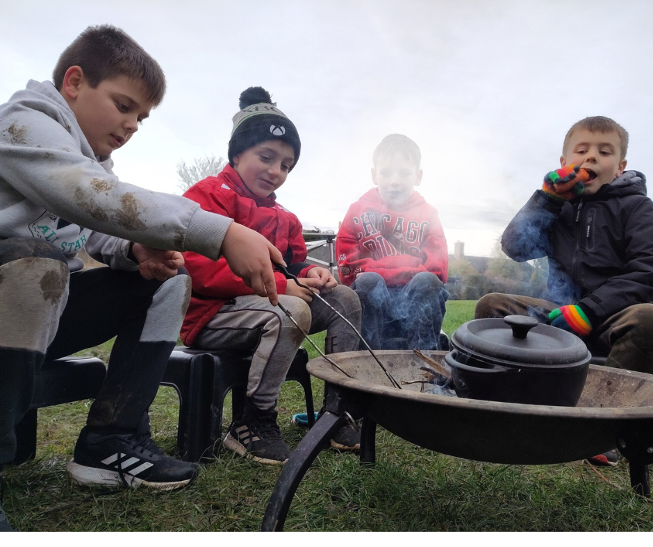 Four young boys sit beside a fire pit, poking it with sticks. Smoke blows up in front of them.  A small Dutch oven pot sits on top of the embers, cooking food outdoors for the youth group to enjoy.