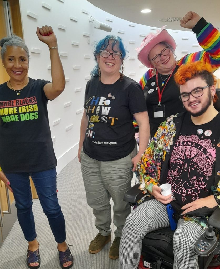 Four people stand in the hall outside a council meeting, where they questioned thecouncil on its socio-economic duty or poverty busting commitments. A black woman on the left has her arm raised in celebration. Her t-shirt reads "More blacks, more Irish, more dogs". Two woman stand beside her, one with a pink cowboy hat, and a person in a wheelchair wearing a t-shirt featuring an illustration of a tree and a slogan which reads "Trans is natural".
