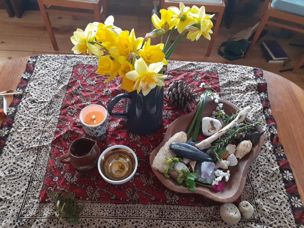 A table set with a cloth, vase of daffodils, a candle, jug of water and small items from nature, a pine cone, stone, sprig of rosemary, a shell. 