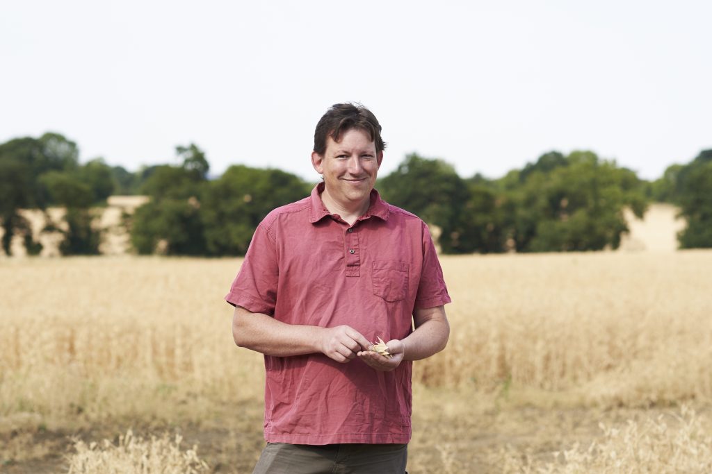 Josiah in a red short-sleeve shirt stands in front of a field of grain. 