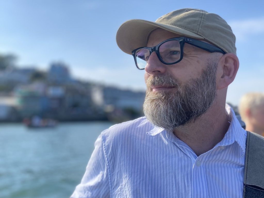 Phil, a man wearing classes and a cap, is looking thoughtfully into the distance, with clear blue water and blue sky behind him. 