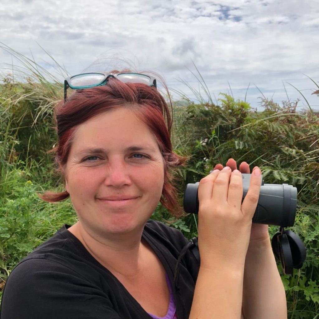 Jess, a woman with red hair, stands in front of a wild hedge holding a pair of binoculars in her hands. 