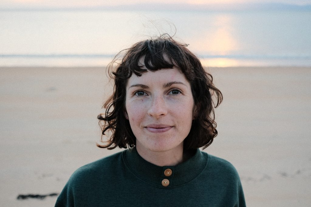 Jo, a woman with dark wavy hair, stands in front of a pale sunset over the sea. 