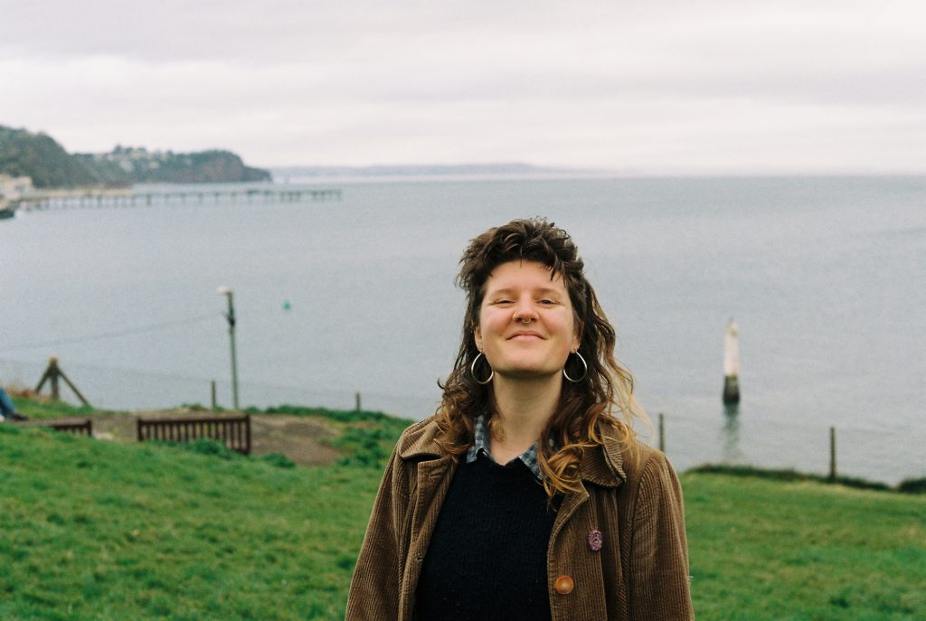 El is pictured in a grassy field overlooking a calm sea with a small pier, a village and headlands in the distance. They have curly brown hair and a smile. 