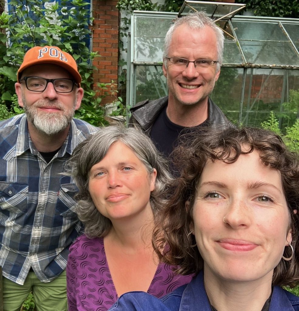 Phil (in an orange hat), Rich (wearing black), Jess (in a purple top) and Joanna (holding camera for a group selfie) stand in front of a red brick building with ferns, shrubs and a weathered greenhouse.